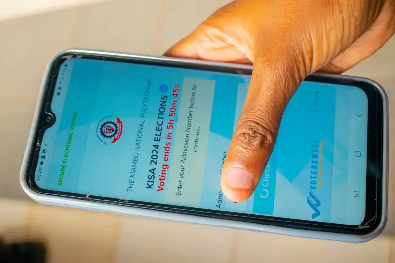 Mkono ukishika simu yenye ukurasa wa kuingia wa Voterswall wa uchaguzi wa 2024 wa Kiambu National Polytechnic na muda uliobaki wa kupiga kura.